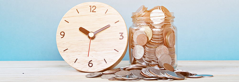 Wooden analog clock next to glass jar overflowing with coins against a blue background.